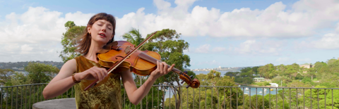 A woman plays the viola and sings in front of a sweeping background of sky, trees, and ocean.